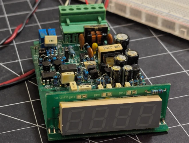 A circuit board with a large LED 7 segment display sits on a desk mat, clamped in a small vice. The LEDs are unlit.