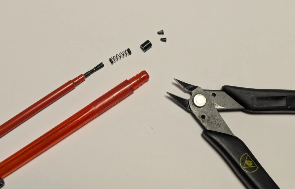A dismantled BIC pen showing the outer plastic tube and a modified inner plastic tube. The inner plastic tube has the spring and black collar detached from the end. A pair of flush cutters rests nearby.
